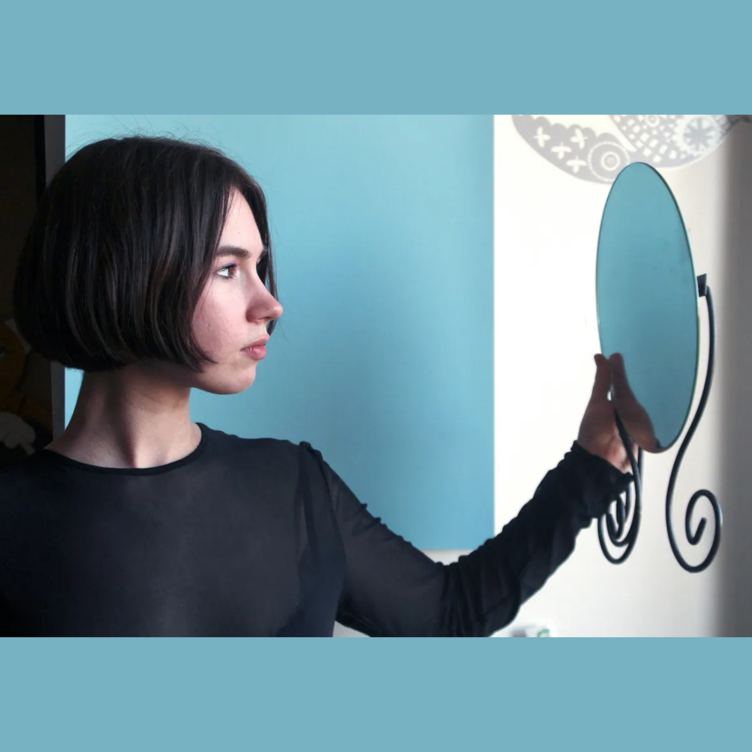 Woman with a short dark bob haircut looking thoughtfully toward her reflection in an oval wall-mounted mirror.